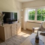 Ein Wohnzimmer mit Fernseher, Sessel und Tisch. Durch ein Fenster blickt man auf Bäume.