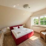 Schlafzimmer mit rotem Bett, Wicker-Sessel und Fenster mit Blick auf Grün.