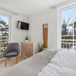 Schlafzimmer mit Bett, Stuhl, Schrank und TV. Große Fenster mit Blick auf Bäume.