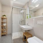 Badezimmer mit Dusche, Waschbecken und Toilette. Holzregal und Heizkörper sichtbar.