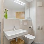 Badezimmer mit Waschbecken, Toilette und Spiegel. Holzstuhl unter dem Waschbecken.