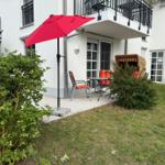 Terrasse mit rotem Sonnenschirm, Tisch und Stühlen. Grünflächen und Hecken umgeben den Außenbereich.