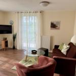 Wohnzimmer mit Sofa, Sessel, Glas-Tisch und Fernseher. Fenster mit weißen Vorhängen.