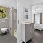 Badezimmer mit Dusche, Toilette und Waschtisch. Im Hintergrund ist ein Schlafzimmer mit Bett und Fenster.