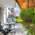Terrasse mit Tisch, drei Stühlen und Sonnenschirm. Hintergrund: Grünflächen und Gebäude.