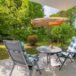 Terrasse mit Tisch, zwei Stühlen und Sonnenschirm. Grünfläche und Bäume im Hintergrund.
