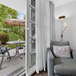 Ein Raum mit grauem Sofa und Stehlampe. Durch die Glastür ist eine Terrasse mit Stuhl und Tisch zu sehen.