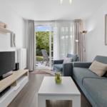 Wohnzimmer mit Sofa, Fernseher und Blick auf Balkon mit Garten.