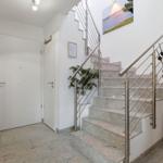 Hallway with stairs, plants, and wall art. White walls and floor tiles.