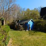Ein blauer Schuppen mit Holzzaun steht auf einer Wiese neben einem Haus.