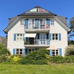 Ein zweistöckiges Haus mit Balkonen und blauen Fensterläden. Umgeben von Grün und Blumen.