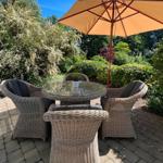 Außenbereich mit Rattan-Sitzgruppe, Glas-Tisch und Sonnenschirm unter blauem Himmel.