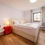 Schlafzimmer mit Bett, zwei Stühlen und rotem Nachttisch. Fenster mit Vorhängen und Wandbild.