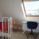 Schlafzimmer mit Schreibtisch, Stuhl und Fenster. Rote Matratze hinter Gitter.