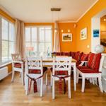 Essbereich mit weißem Tisch, Stühlen und Sitzbank mit roten Kissen. Große Fenster, Holzboden und orange Wände.