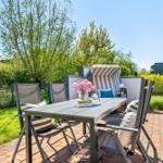 Terrasse mit Tisch, Stühlen und Strandkorb. Grünfläche und Zaun im Hintergrund.