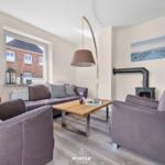 Wohnzimmer mit Sofa, zwei Sesseln, Kamin und Fernseher. Fenster zeigt Haus außen.