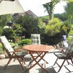 Terrasse mit Tisch, Stühlen und Sonnenschirm. Hintergrund: Garten mit Sträuchern und Haus.