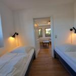 Zwei Betten mit weißen Bettwäsche, Wandlampen und Holzboden. Durchgang zu Wohnzimmer mit Tisch.