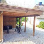 Holzüberdachung mit Fahrrädern und Schuppen neben einem Strohdachhaus.