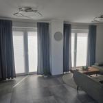 Wohnzimmer mit grauen Möbeln, Fenstern und Teppich. Deckenleuchten und Wanduhr sichtbar.