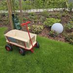 Ein roter Kinderwagen steht auf dem Rasen neben einem Beet mit Pflanzen und einem Steinball.