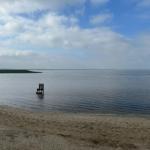 Eine Stuhl am Strand im Wasser, Blick auf ruhiges Gewässer und Himmel mit Wolken.