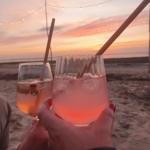 Zwei Gläser mit Getränken auf dem Strand bei Sonnenuntergang mit Lichterkette im Hintergrund.