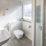 Badezimmer mit Waschbecken, Toilette und Dusche. Fenster mit Pflanze und Handtuchhalter.