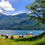 Grüner Strand mit Bäumen, Menschen und einem See vor Bergen.