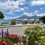 Terrasse mit Sonnenschirmen am See, umgeben von Blumen und Bergen.