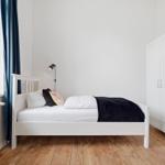 A bedroom with a white bed, blue curtains, and wooden floor. A white wardrobe is on the wall.
