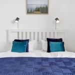 A bed with blue quilt and pillows, white wall, two wall lamps, and a picture above the headboard.