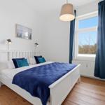 Bedroom with white bed, blue quilt, and wooden floor. Window with blue curtains.