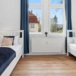 Two single beds with white linens and dark blankets stand before a window with blue curtains.