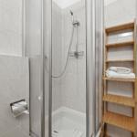 Shower with glass enclosure, wooden shelf, and toilet paper holder on wall.