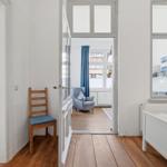 Bedroom with wooden floor and white bed frame. Door leads to living area with armchair.