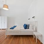 Schlafzimmer mit weißem Bett, Holzboden und Spiegel. An der Wand hängen zwei Lampen.