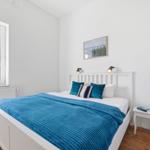 A bedroom with a white bed, blue blanket, and window with view of trees.