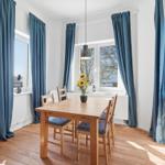 Dining room with wooden table, chairs, and blue curtains. Sunflowers on table. Wooden floor.