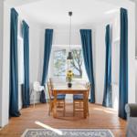 Dining area with wooden table, blue curtains, and sunflowers. Wooden floor and sofa in foreground.