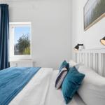 Bedroom with white bed, blue blanket, and wall lamp. Window with blue curtains.
