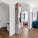 Badezimmer mit Dusche, Toilette und Waschbecken. Wohnzimmer mit Sofa und Schlafzimmer im Hintergrund.