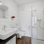 Badezimmer mit Dusche, Waschbecken und Toilette. Weiße Fliesen und Holzboden.