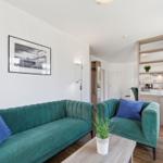 Ein Wohnzimmer mit grünem Sofa, blauen Kissen und einem Holztisch. Im Hintergrund ist die Küche sichtbar.