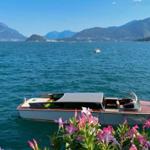 Ein Boot liegt vor einer Bucht mit Bergen im Hintergrund und rosa Blumen im Vordergrund.