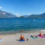 Strand mit Steinen, Menschen baden und entspannen, Berge im Hintergrund, Boot auf dem Wasser.