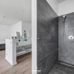 Dusche mit grauen Fliesen, Waschbecken und Schlafzimmer im Hintergrund.