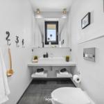 Badezimmer mit Waschtisch, Spiegel und Toilette. An der Wand hängen Handtücher und ein Bürstenhalter.