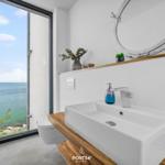 Badezimmer mit Blick aufs Meer, Waschbecken auf Holz, runder Spiegel, Pflanzen und Toilette.